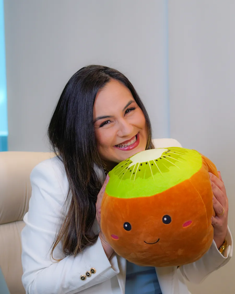 Juliana Araújo sorrindo e abraçando uma almofada em forma de kiwi